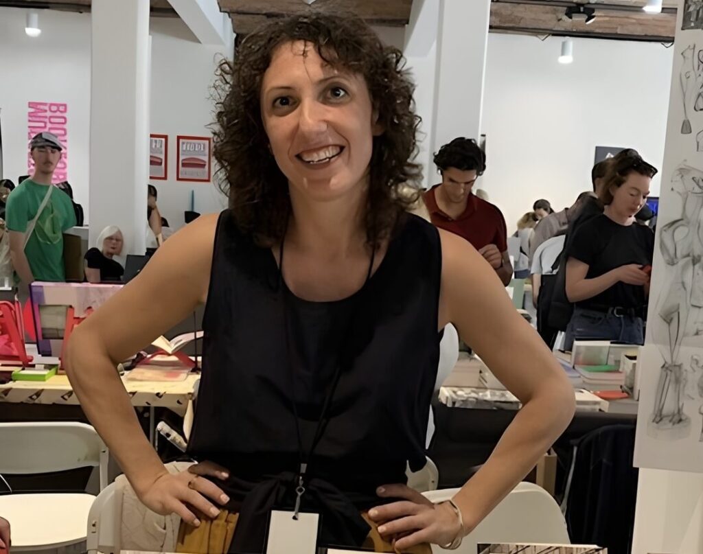 Image: Julia Klein, wearing a black tank top and smiling for the camera, poses at her booth at the New York Art Book Fair. Photo by Lisa Pearson.
